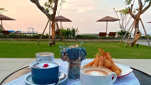 A table with a dessert, two cups of coffee, and a plate of fried food, set outdoors with umbrellas and a view of the sea. - Putri Duyung Ancol (Jakarta)