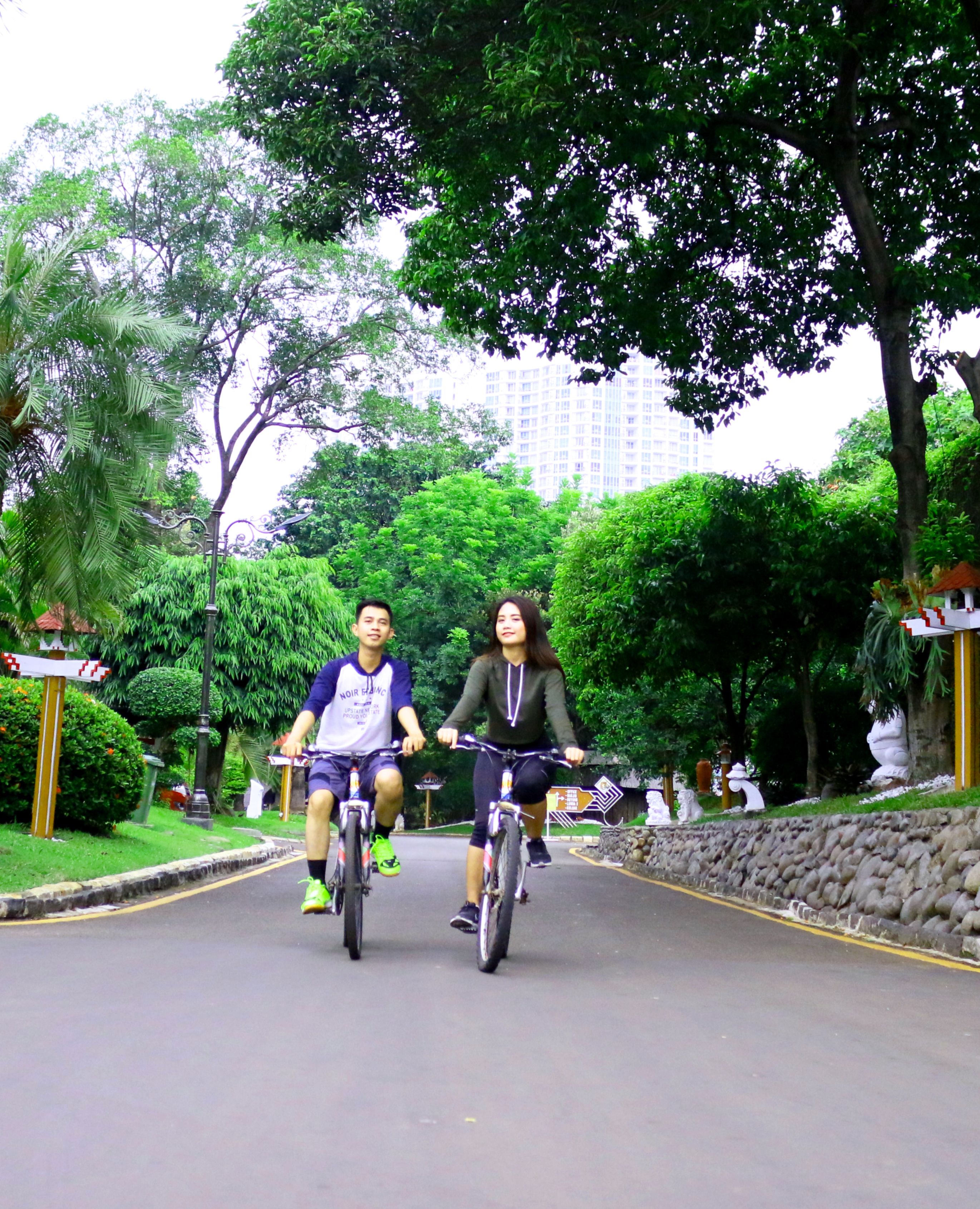 Two people riding bicycles on a paved path surrounded by greenery and trees.