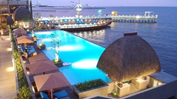A poolside area with lounge chairs and umbrellas, a thatched roof structure, and a view of the sea and a pier with boats. - Putri Duyung Ancol (Jakarta)
