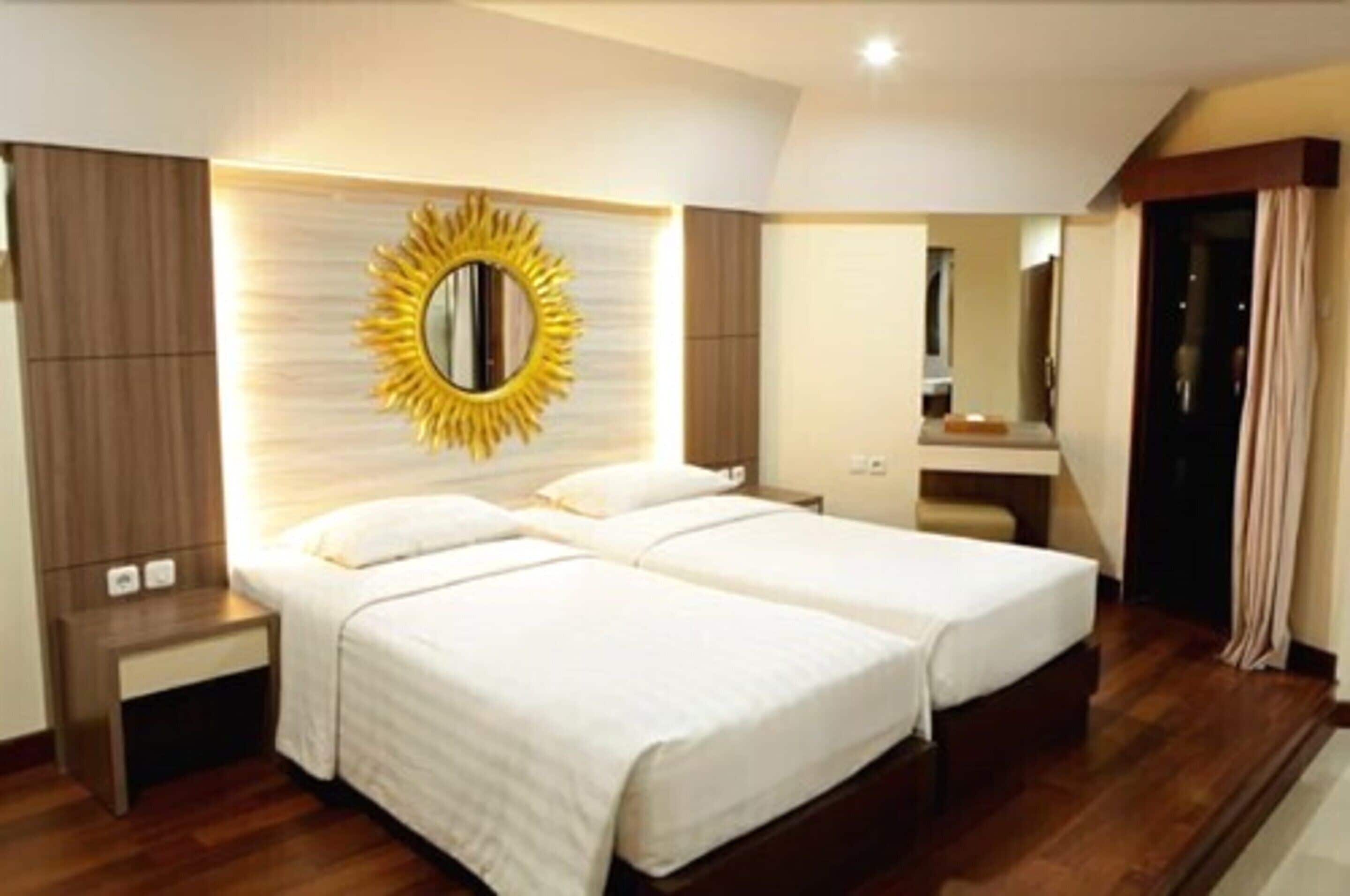 A hotel room with two beds, a large mirror with a sunburst frame, wooden paneling, and a small bedside table.
