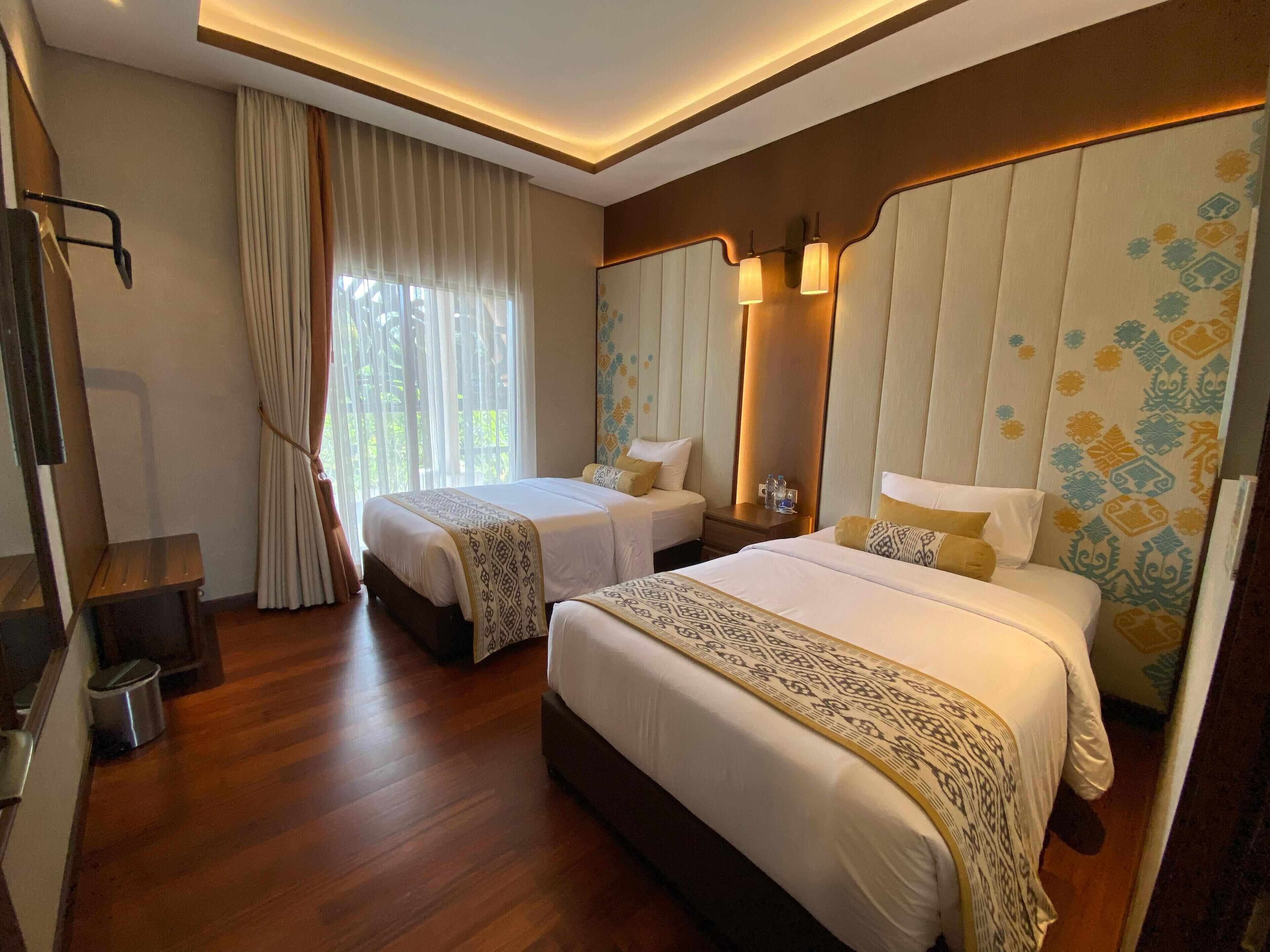 A hotel room with two beds, wooden flooring, a large mirror, and a window with curtains.