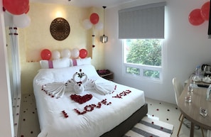 Habitación de hotel impecable, con una cama decorada con pétalos de rosa y toallas con forma de cisne, un rincón de comedor con una mesa dispuesta para dos personas, y una ventana que ofrece vistas a zonas verdes.
