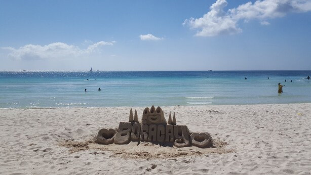 A sandy beach with a sandcastle in the foreground and a clear blue sea in the background.