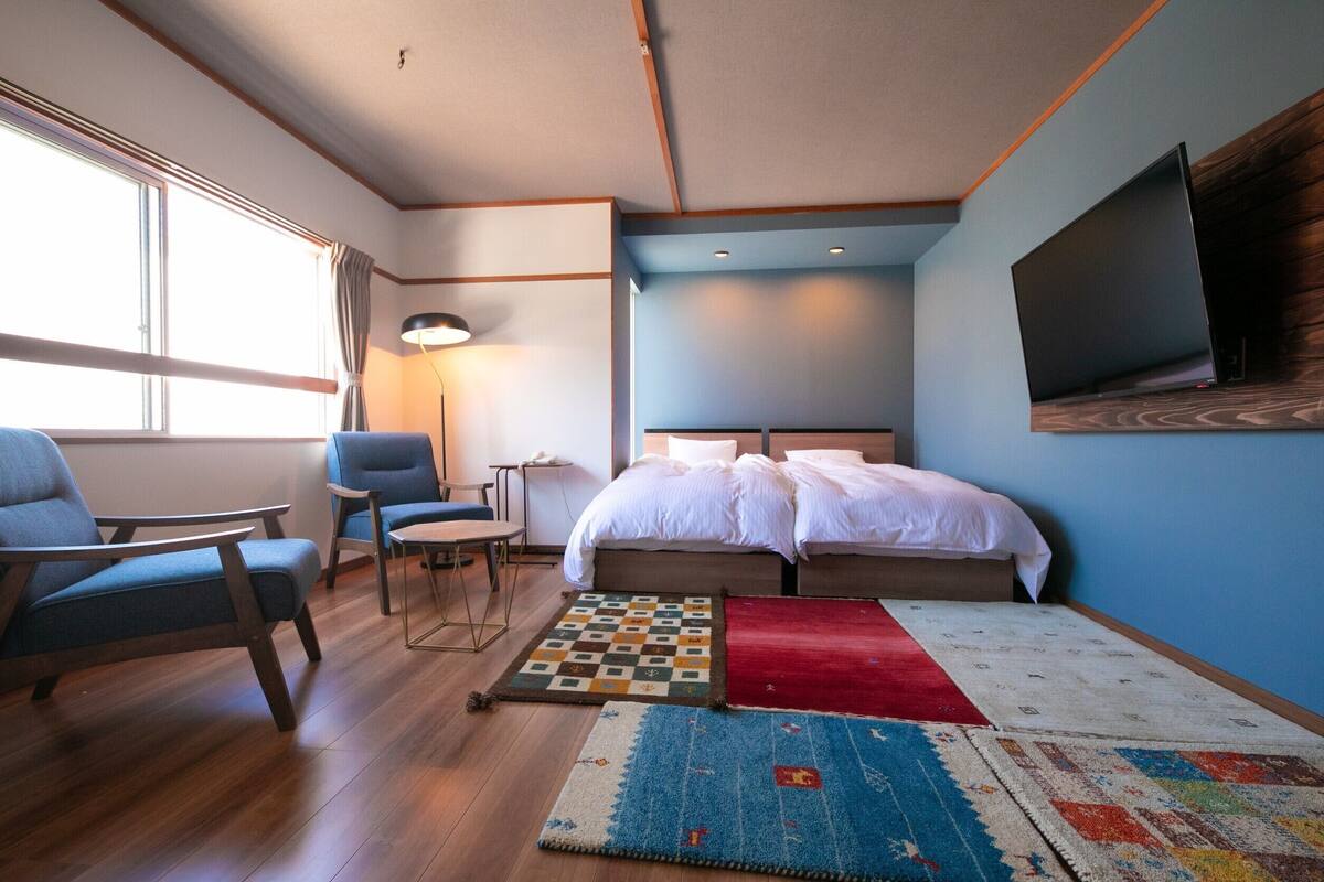 Una habitación de hotel con una cama, dos sillas, un escritorio, una lámpara y un televisor montado en la pared.