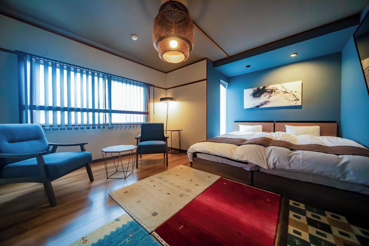 Una habitación de hotel moderna con una cama, dos sillas, una mesita pequeña y un ventanal grande con persianas.