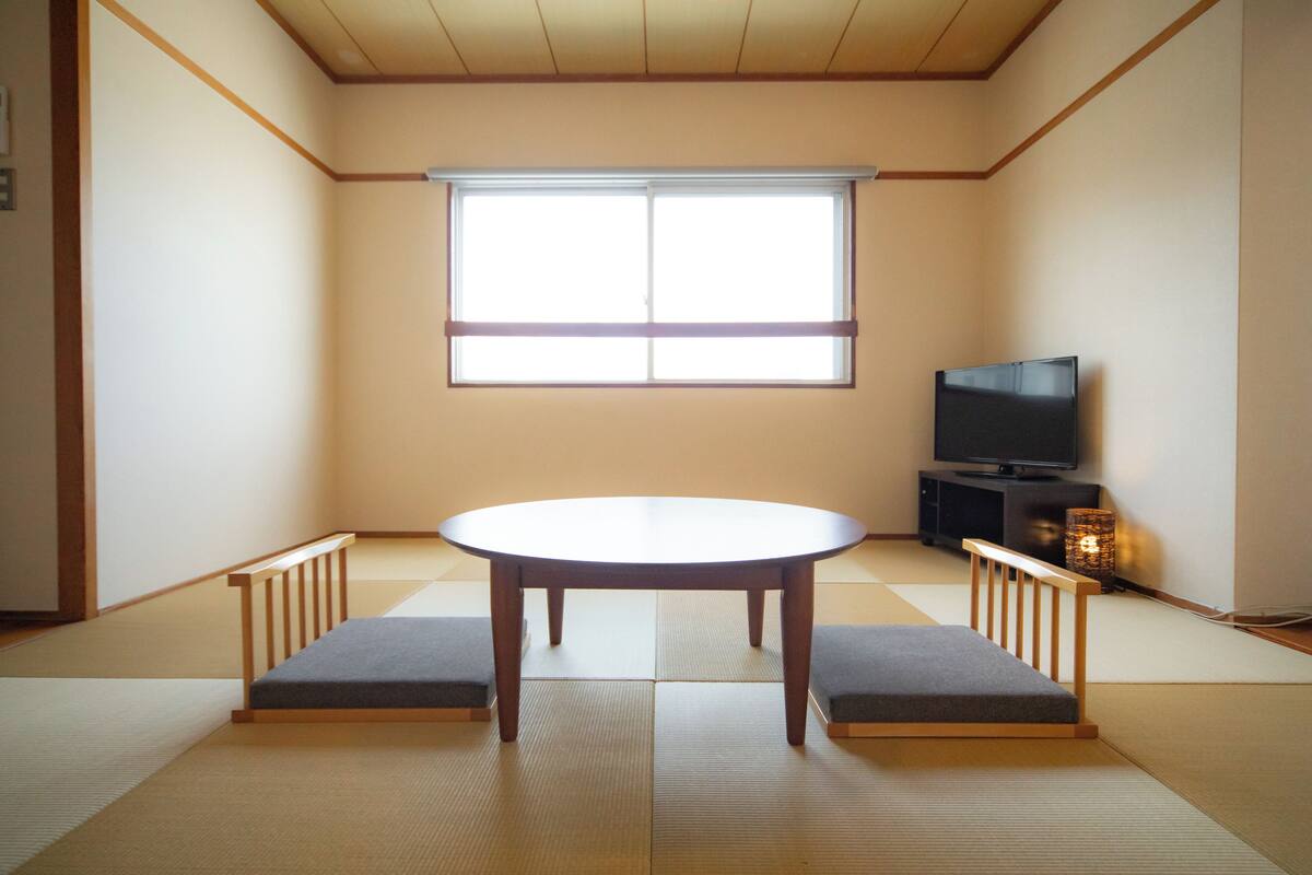 Una habitación con suelo de tatami, una mesa baja, cojines, un televisor y una ventana.