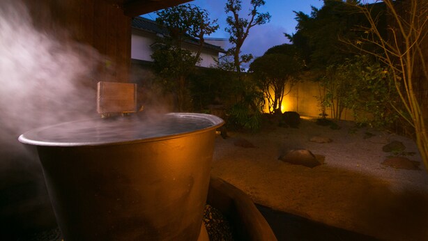 A large metal pot emitting steam, placed outdoors under a wooden structure with a garden and trees in the background. - Kunisakiso (Beppu)