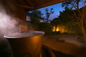 A large metal pot emitting steam, placed outdoors under a wooden structure with a garden and trees in the background.