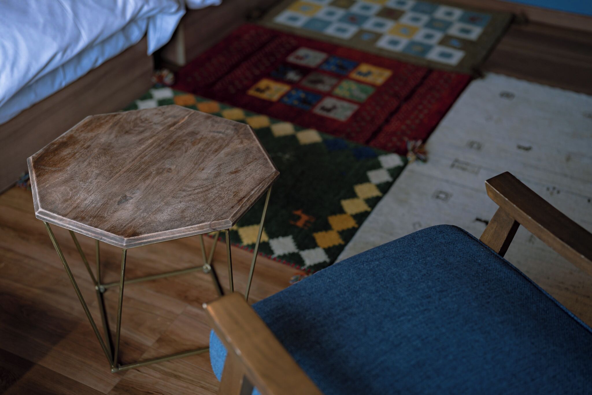 Una habitación con un taburete de madera de ocho lados, una silla azul acolchada, un tapiz acolchado y una cama con ropa de cama blanca.