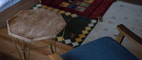Una habitación con un taburete de madera de ocho lados, una silla azul acolchada, un tapiz acolchado y una cama con ropa de cama blanca.