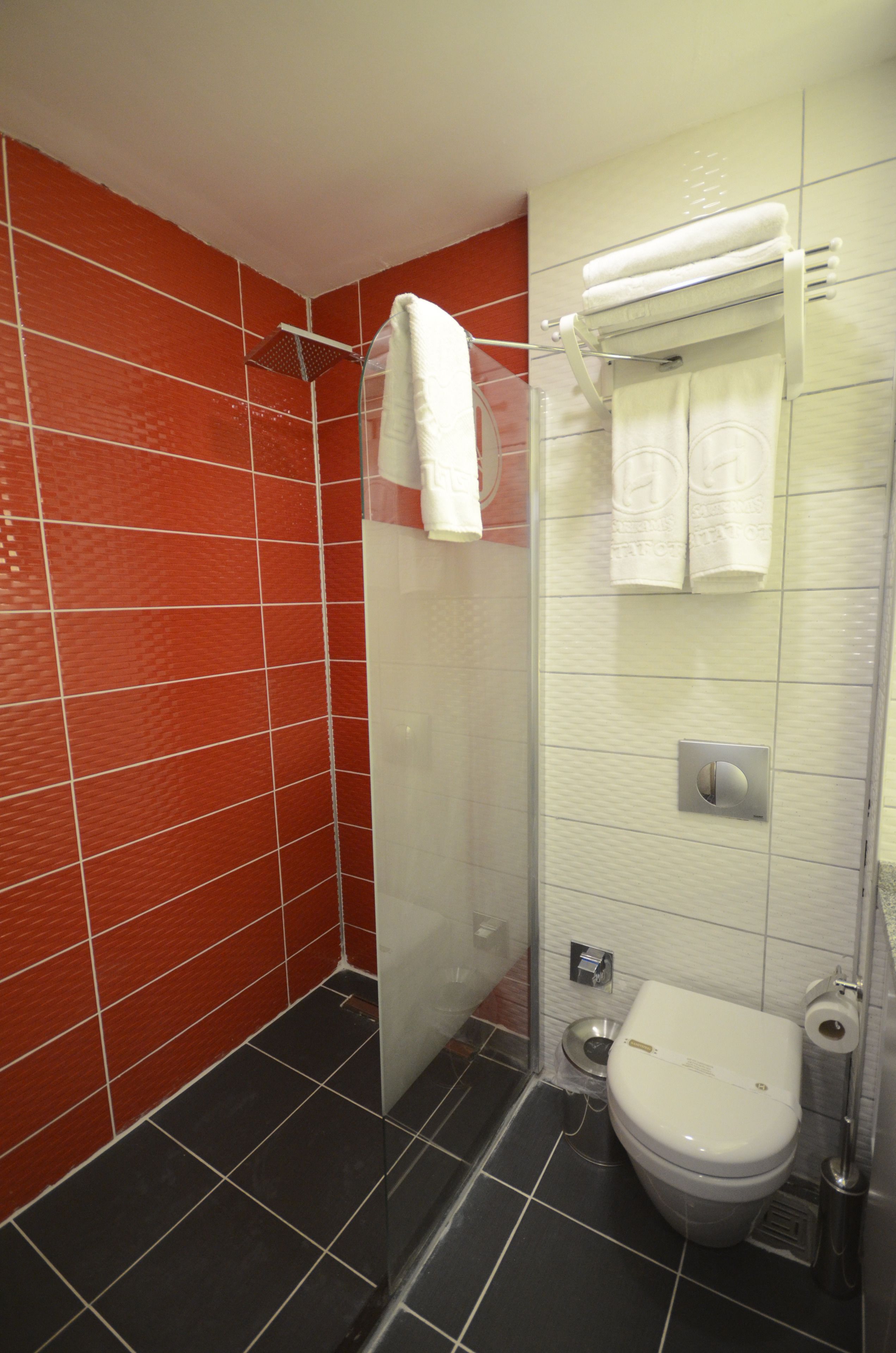 Een badkamer met rode betegelde wanden, een wit toilet, een glazen douchecabine en een handdoekrek met handdoeken.