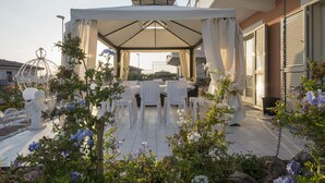 En udendørs terrasse med hvide borde og stole, omgivet af planter og blomster.