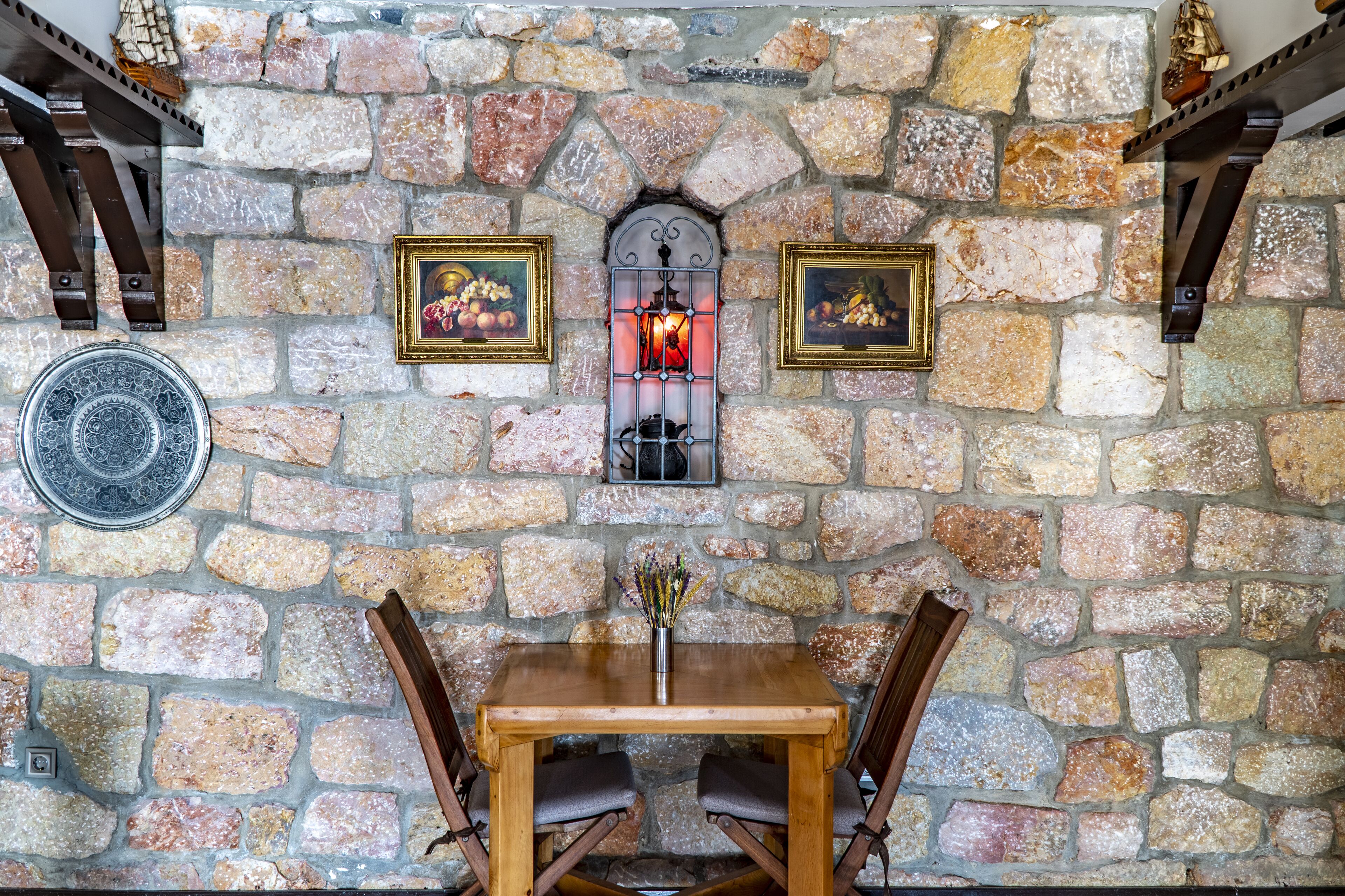 A stone wall with framed artwork, a table with chairs, and a decorative lantern.