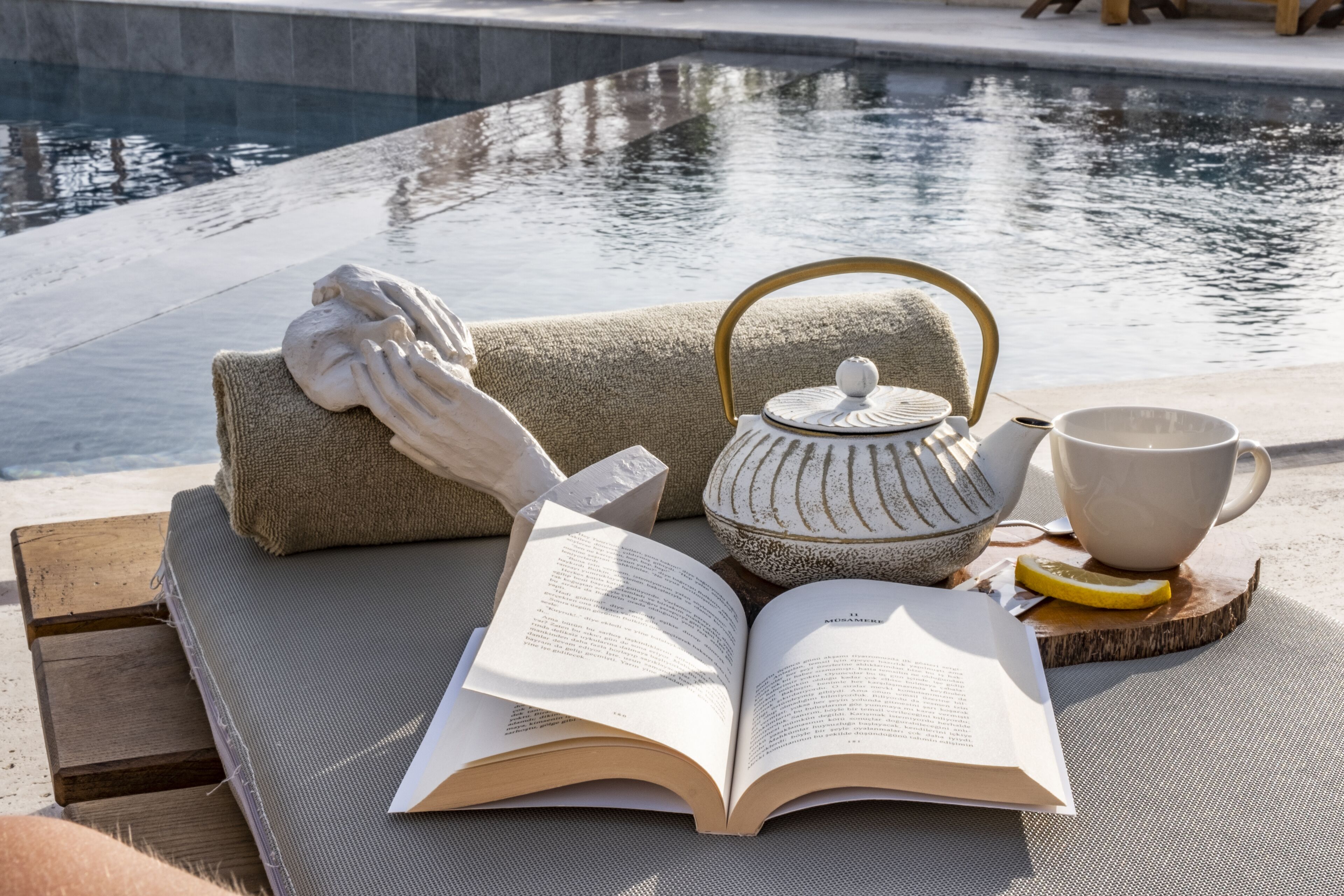 A book, a teapot, a cup, and a lemon on a wooden table by a pool.