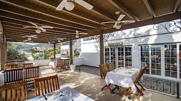 Un área de comedor al aire libre con mesas y sillas de madera, ventiladores de techo y grandes ventanales que ofrecen vistas de la ciudad.