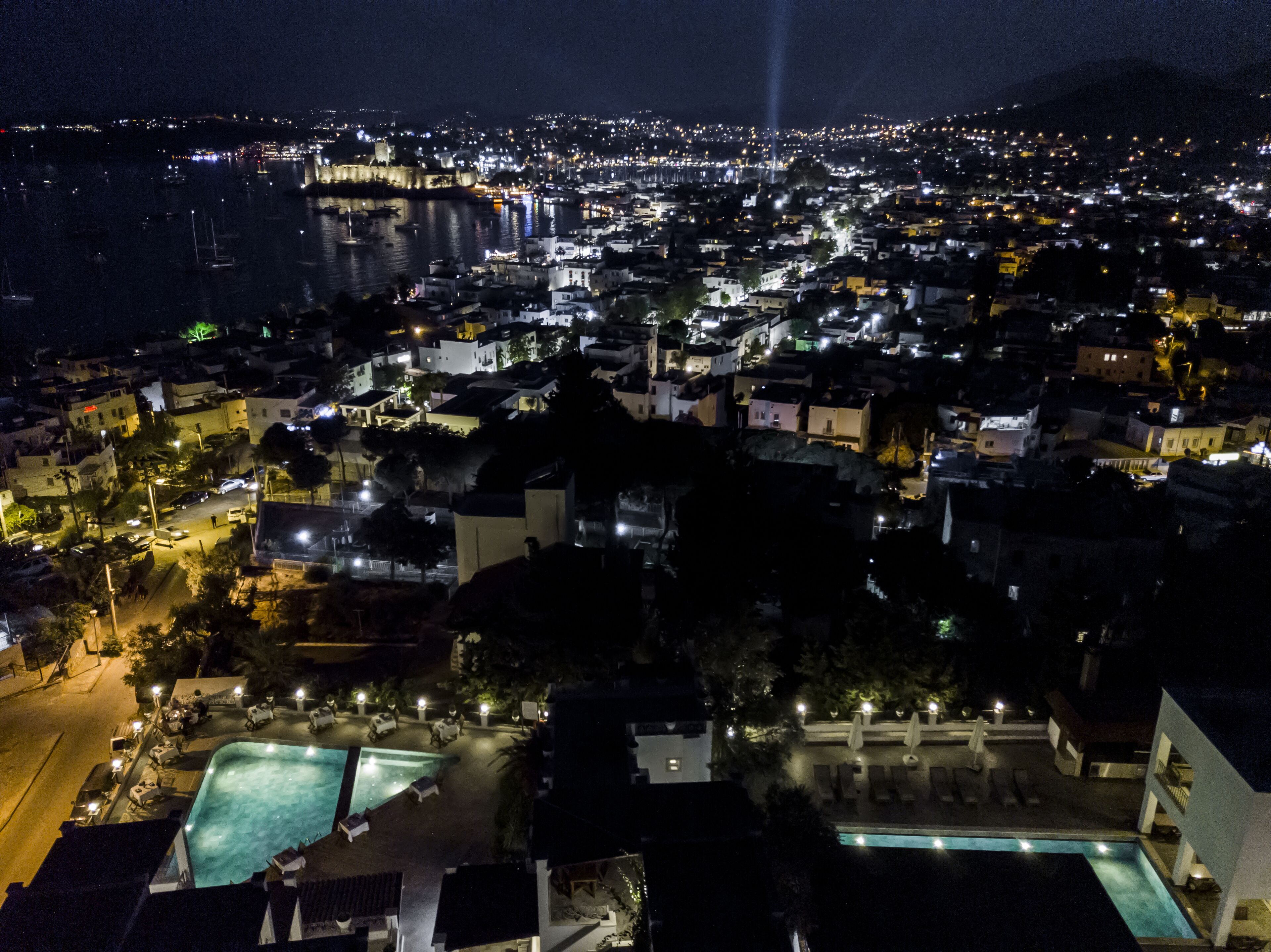 A nighttime aerial view of a coastal city with a prominent castle-like structure, a marina, and a well-lit swimming area with lounge chairs.