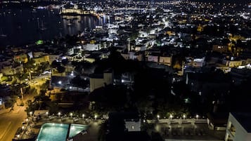 A nighttime aerial view of a coastal city with a prominent castle-like structure, a marina, and a well-lit swimming area with lounge chairs.