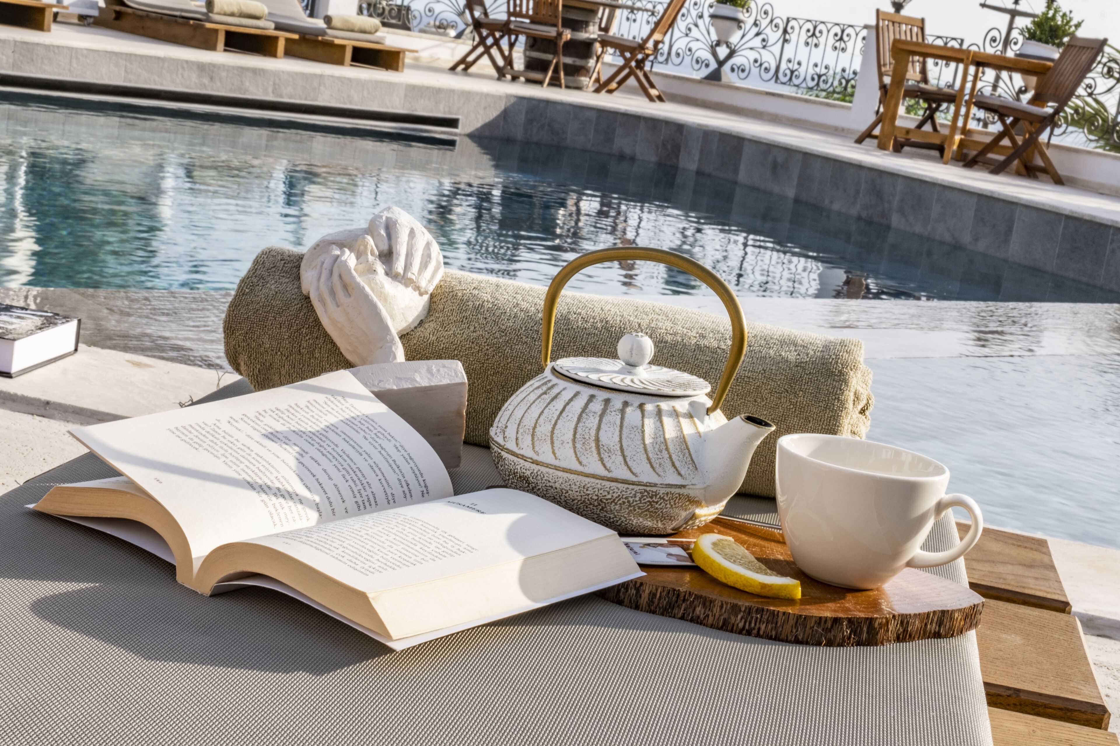 A poolside setting with a book, teapot, mug, and lemon slice on a wooden tray.