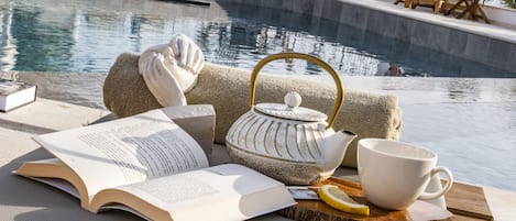 A poolside setting with a book, teapot, mug, and lemon slice on a wooden tray.