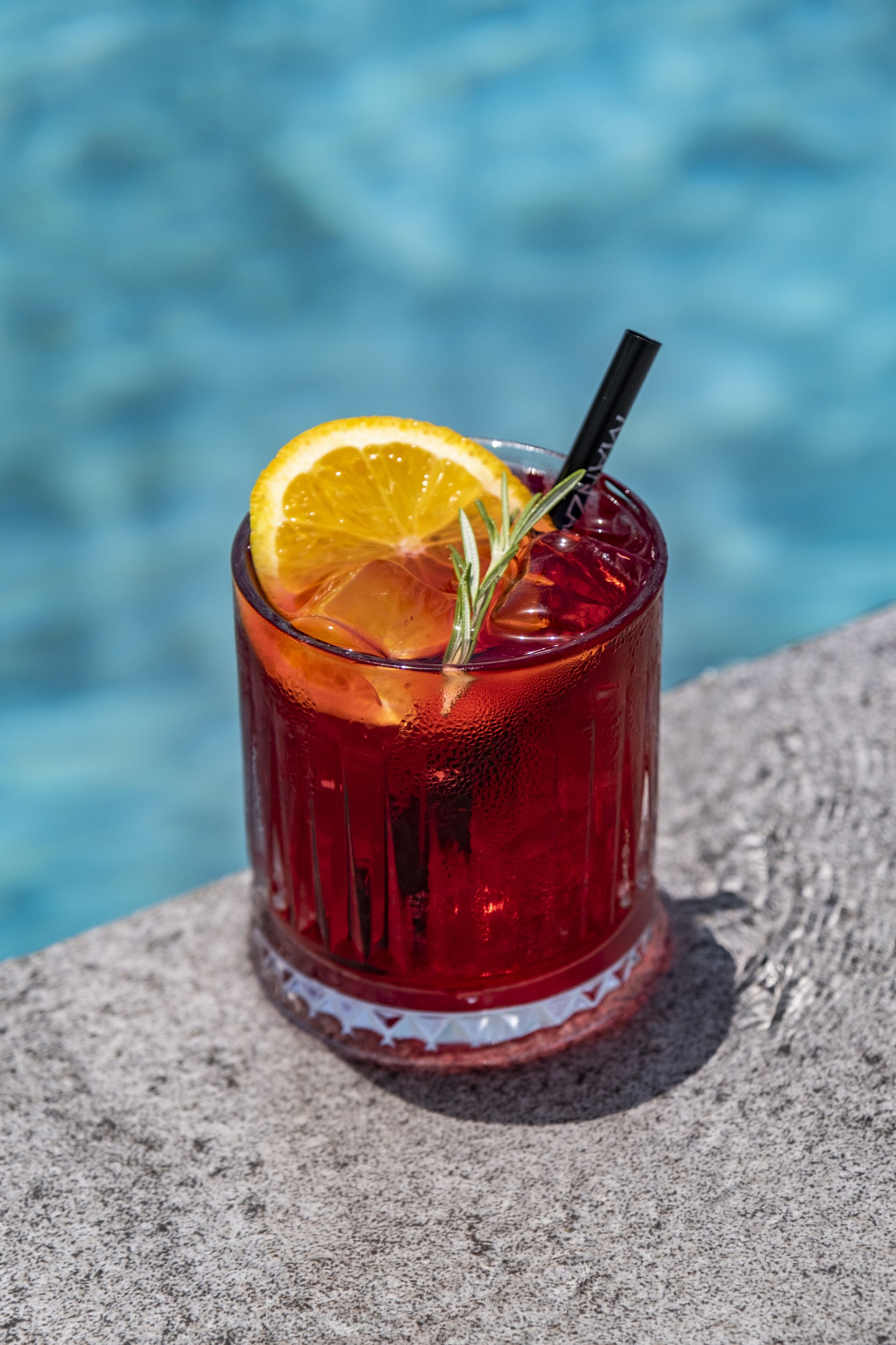 A glass of red drink with a lemon slice and a straw, placed on a stone surface near a body of water.