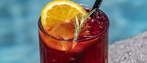 A glass of red drink with a lemon slice and a straw, placed on a stone surface near a body of water.