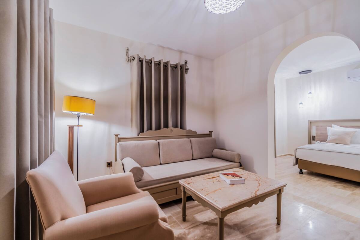 Habitación de hotel con sofá, una silla, una mesita, cama y ventana con cortinas.