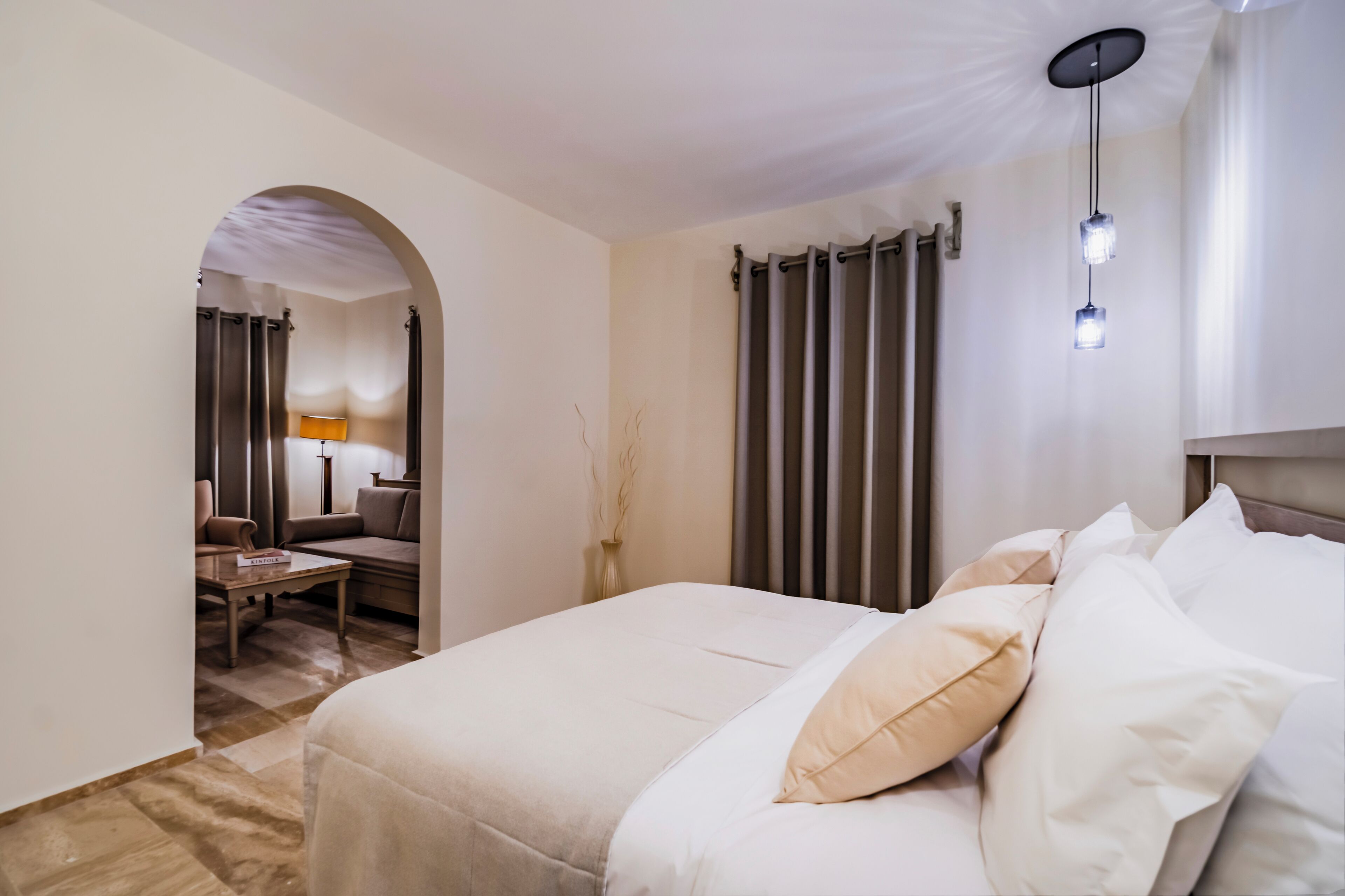 Ein Hotelzimmer mit einem großen Bett, einem Schreibtisch und einem Sessel. Auf dem Schreibtisch steht eine Lampe und am Fenster ein Vorhang.