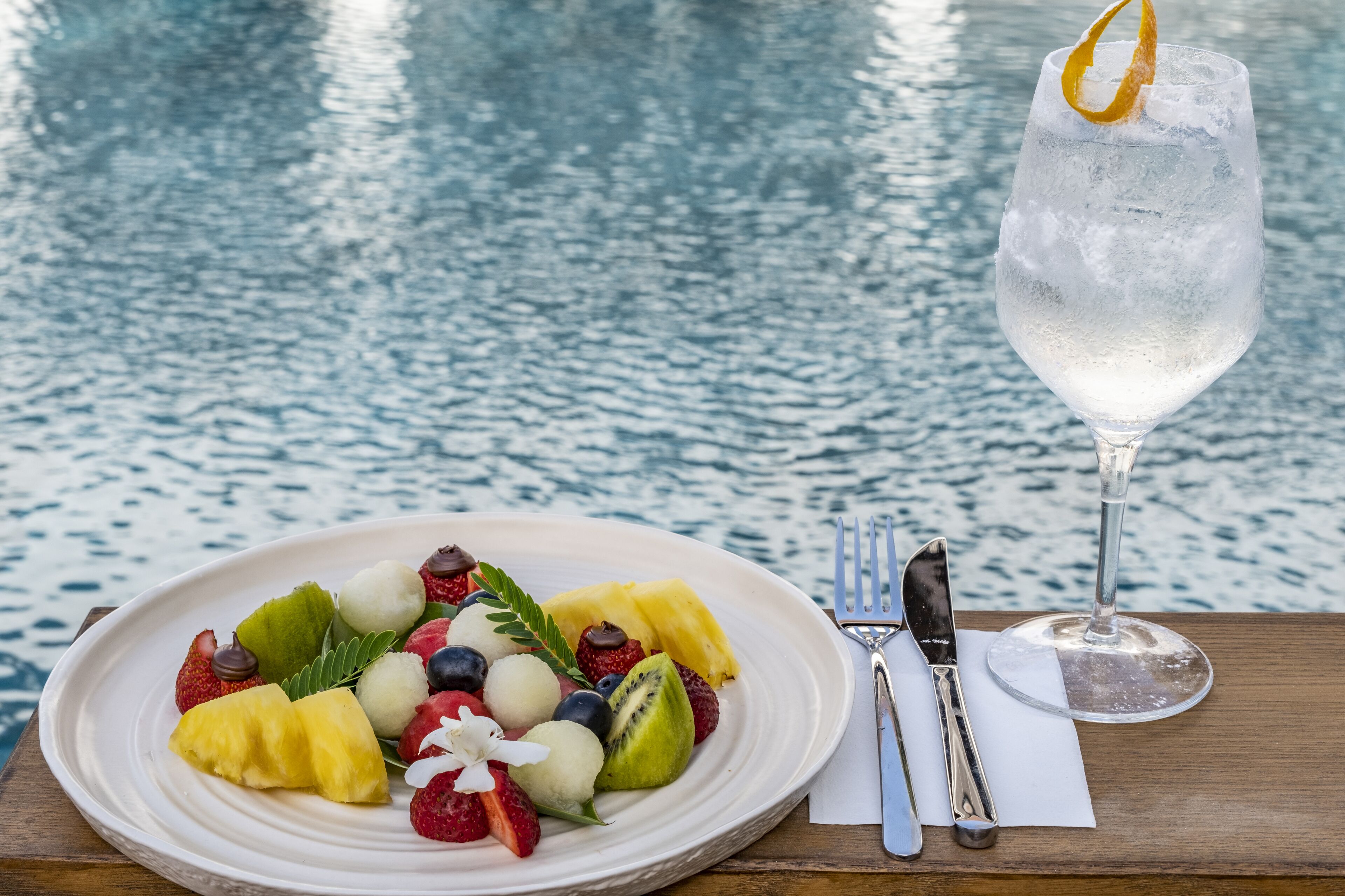 A plate of fruit salad with a glass of water and cutlery on a wooden table by a body of water.