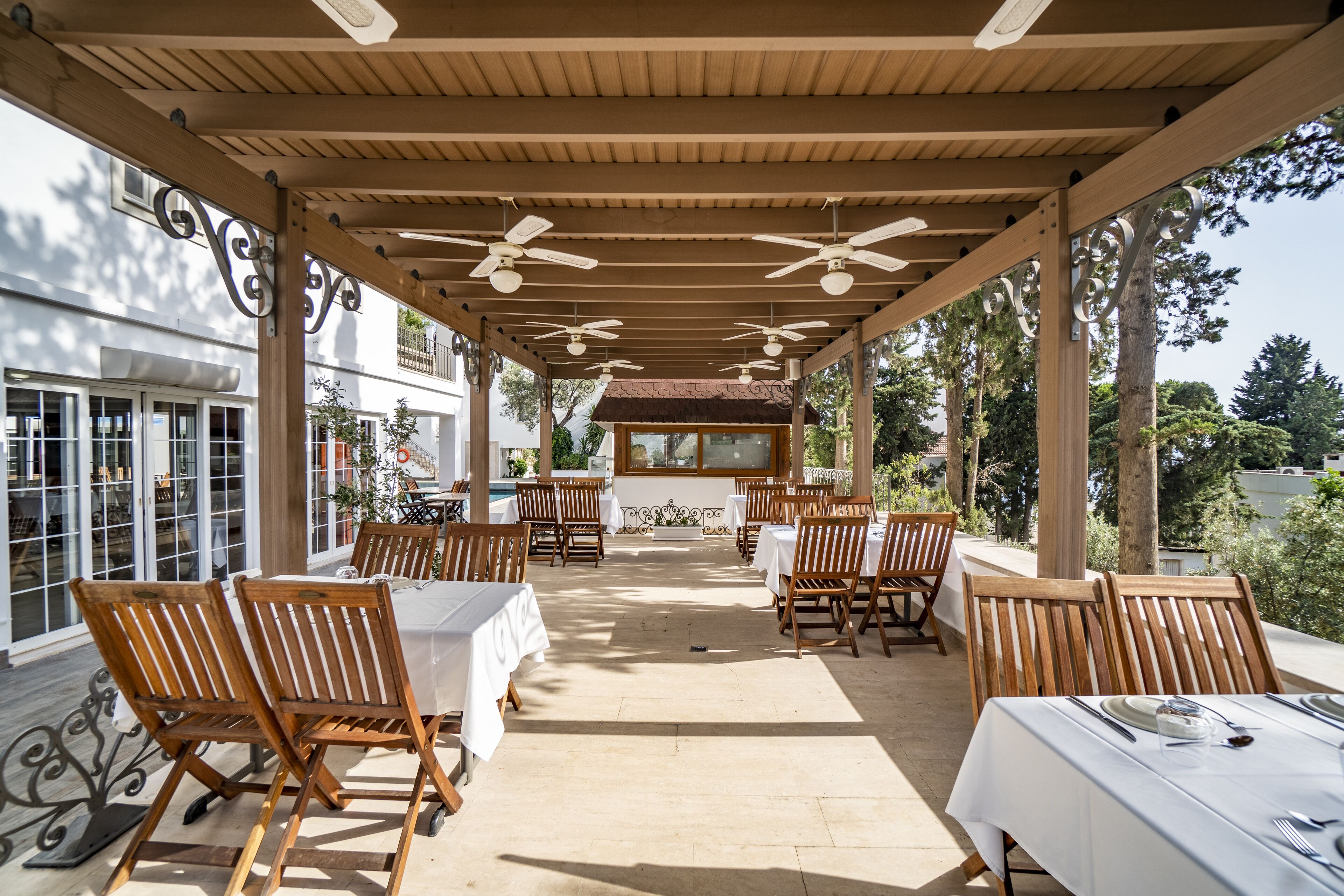 An outdoor patio with wooden tables and chairs, ceiling fans, and a view of the surrounding area.
