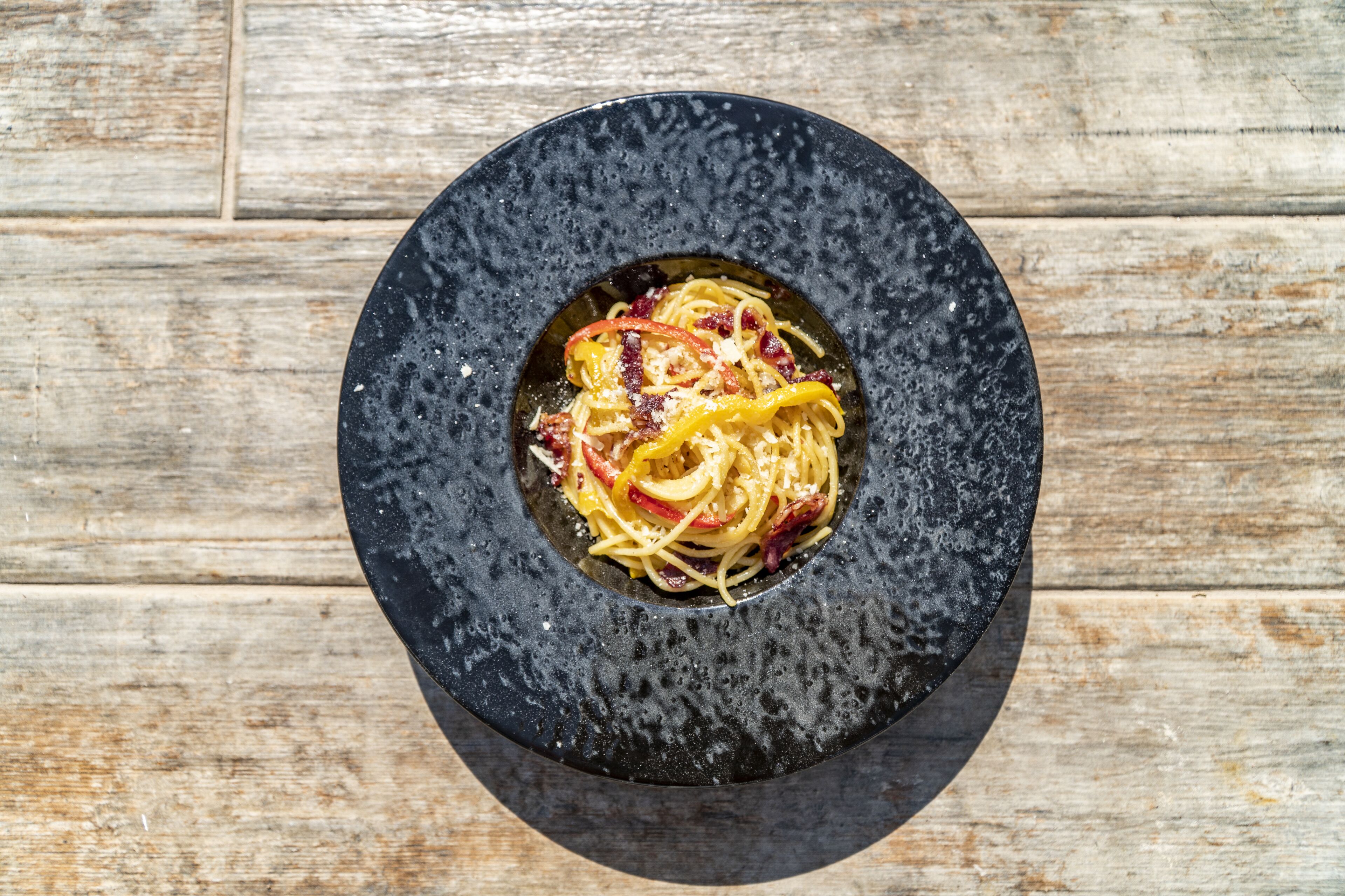 Un bol noir contenant des spaghettis et une tranche de poivron jaune, le tout posé sur une surface en bois.