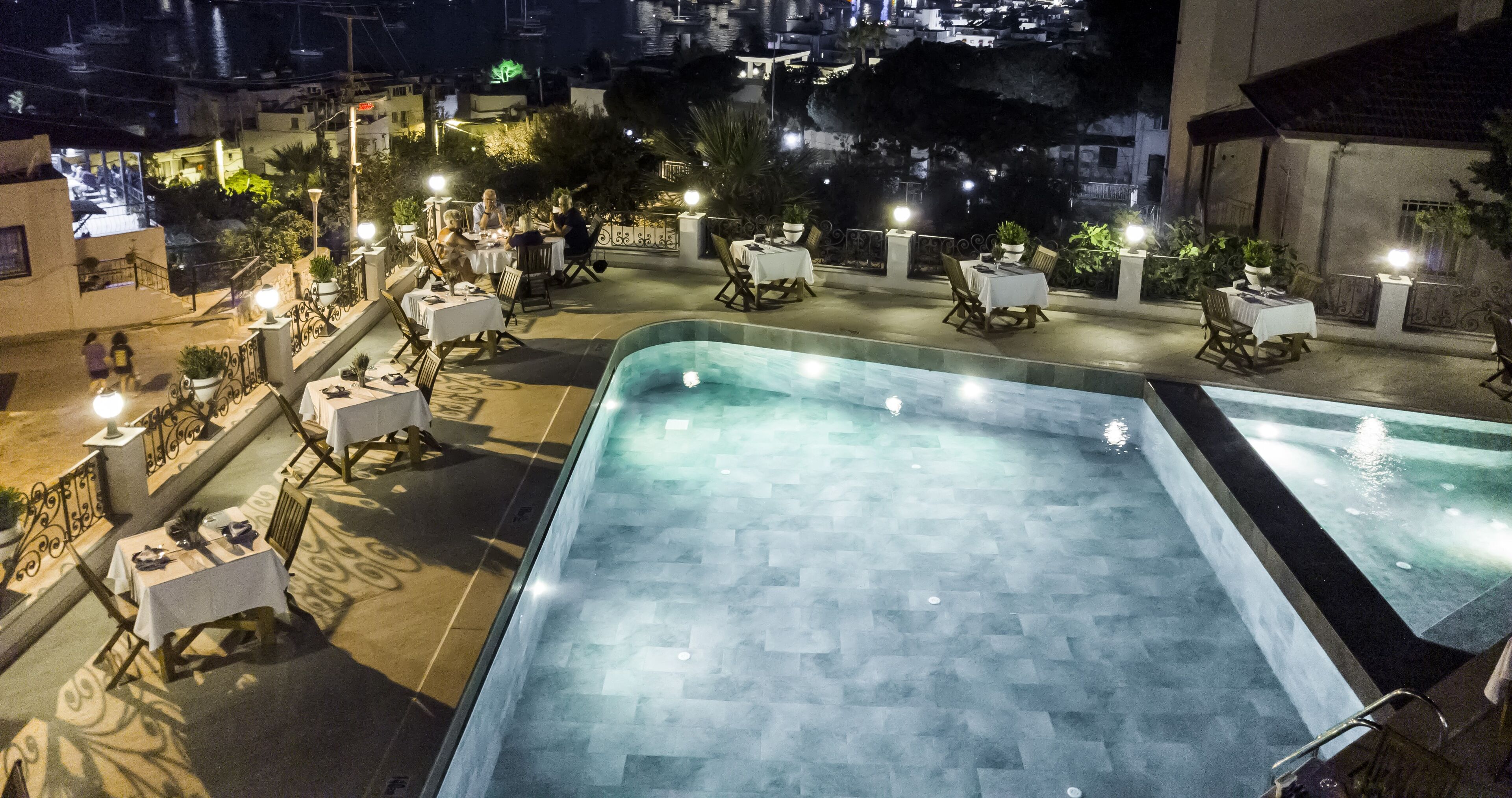 Un área de piscina en la azotea con varias mesas y sillas, rodeada de plantas en macetas e iluminada con luz ambiental.