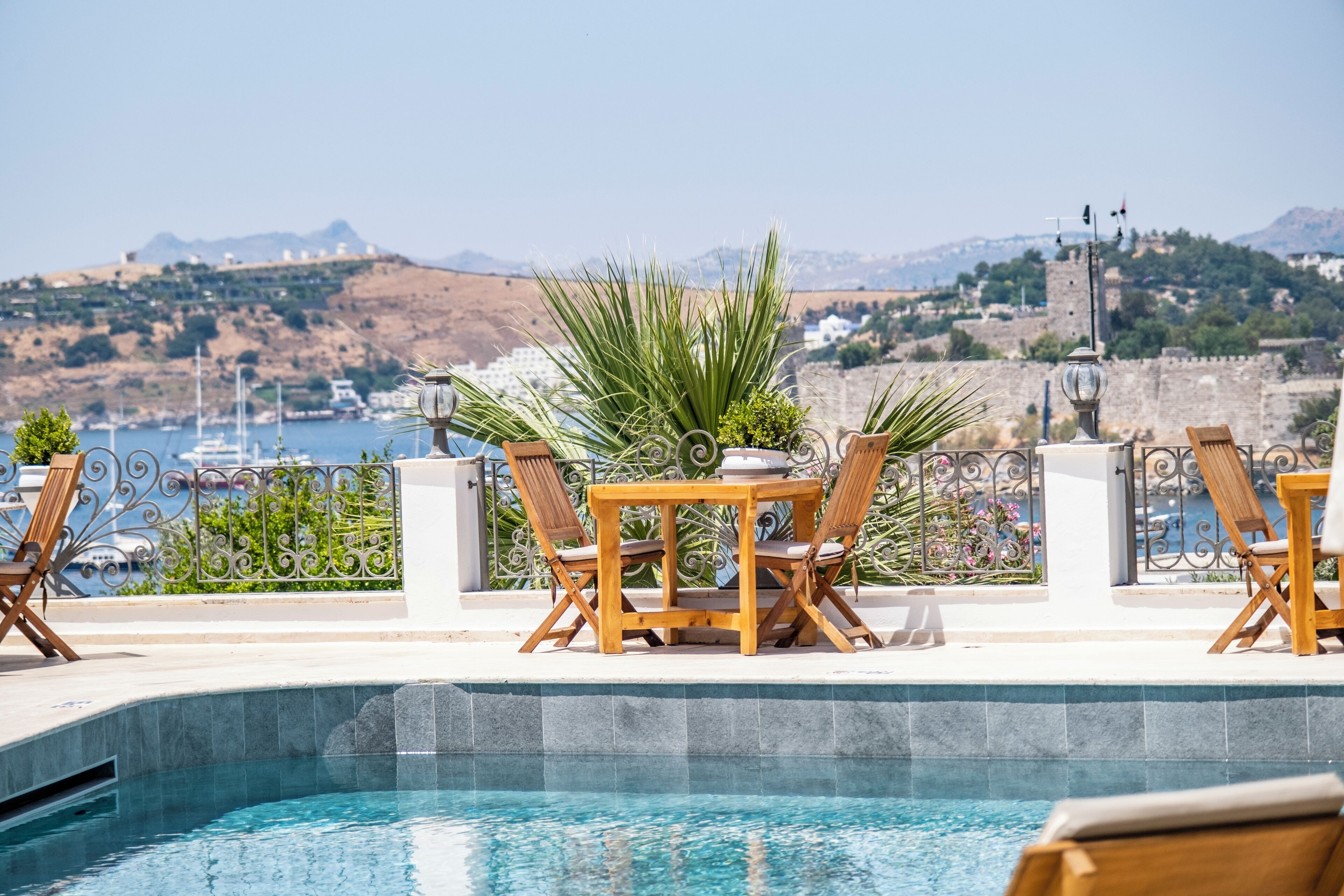 A poolside area with a view of the sea and hills, wooden chairs, a table, and a potted plant.