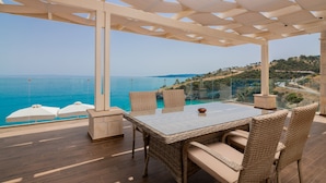 A terrace with a glass balustrade overlooking a coastal view, featuring wicker furniture and a clear sky. - Orient Villas (Zakynthos)
