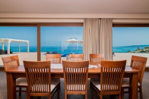 A dining area with wooden chairs and tables, large windows with a view of the ocean and beach. - Orient Villas (Zakynthos)