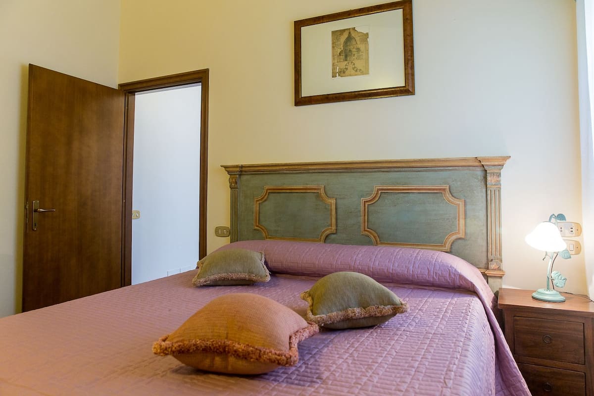 Un dormitorio con cabecera de madera, una cama con colcha rosa y dos almohadas. Hay una mesita de noche con una lámpara y un cuadro en la pared.