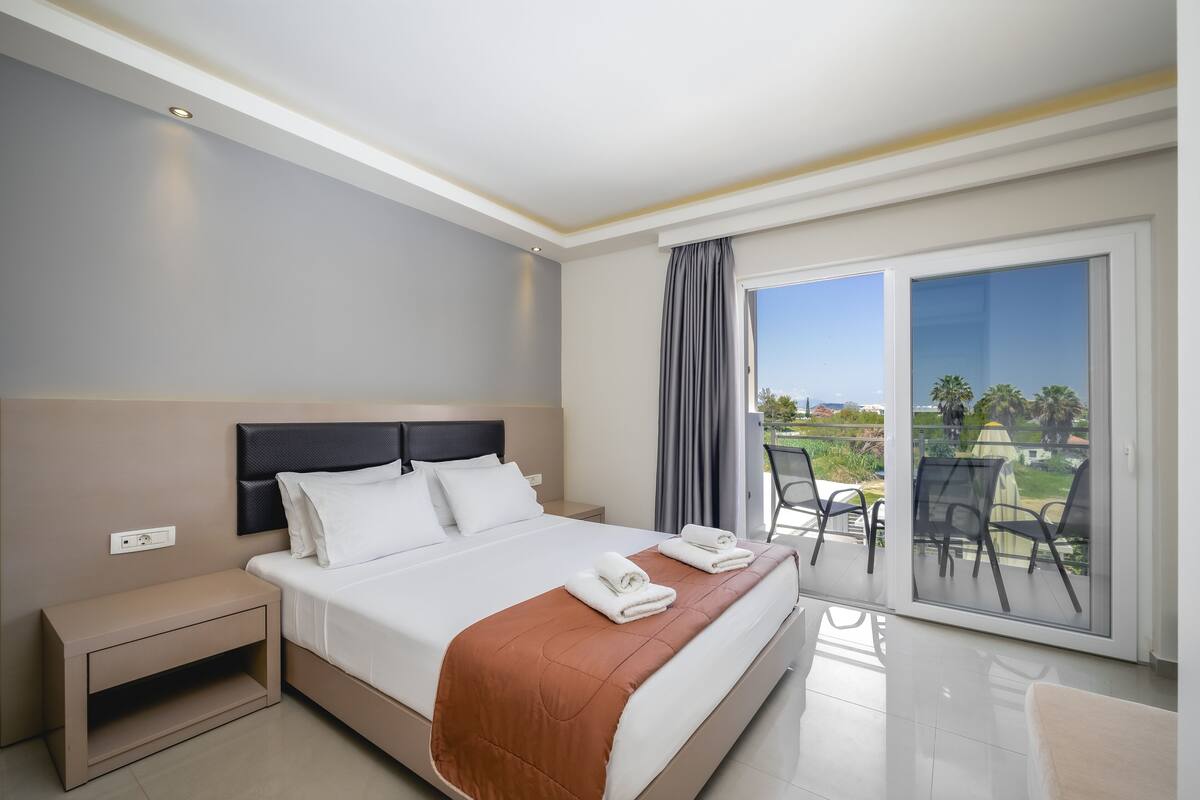 Habitación de hotel moderna con una cama grande, una mesita de noche, un balcón con sillas y vistas al exterior.