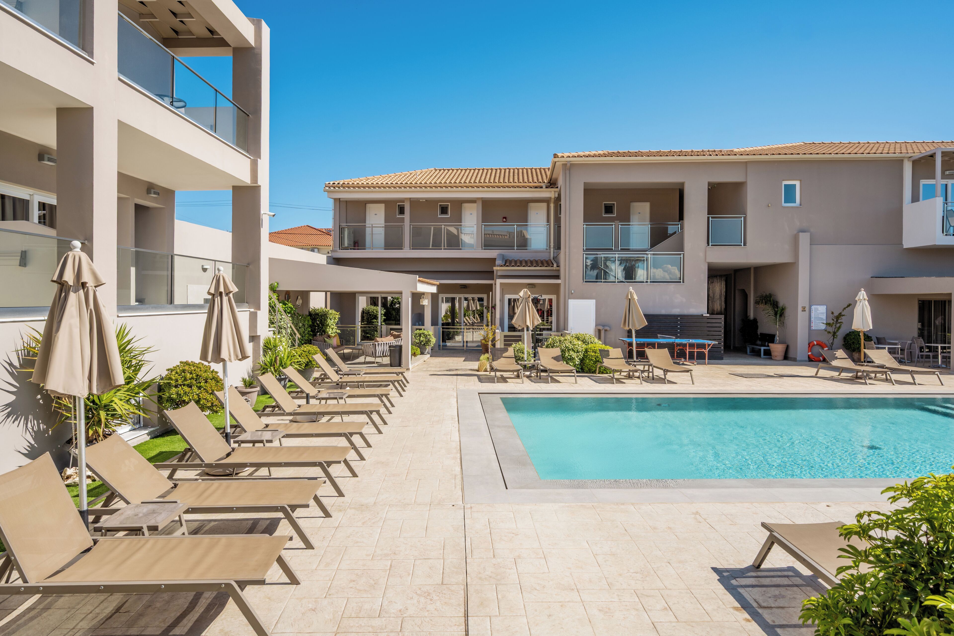 A pool area with lounge chairs and umbrellas, a clear blue sky, and a modern building with balconies.
