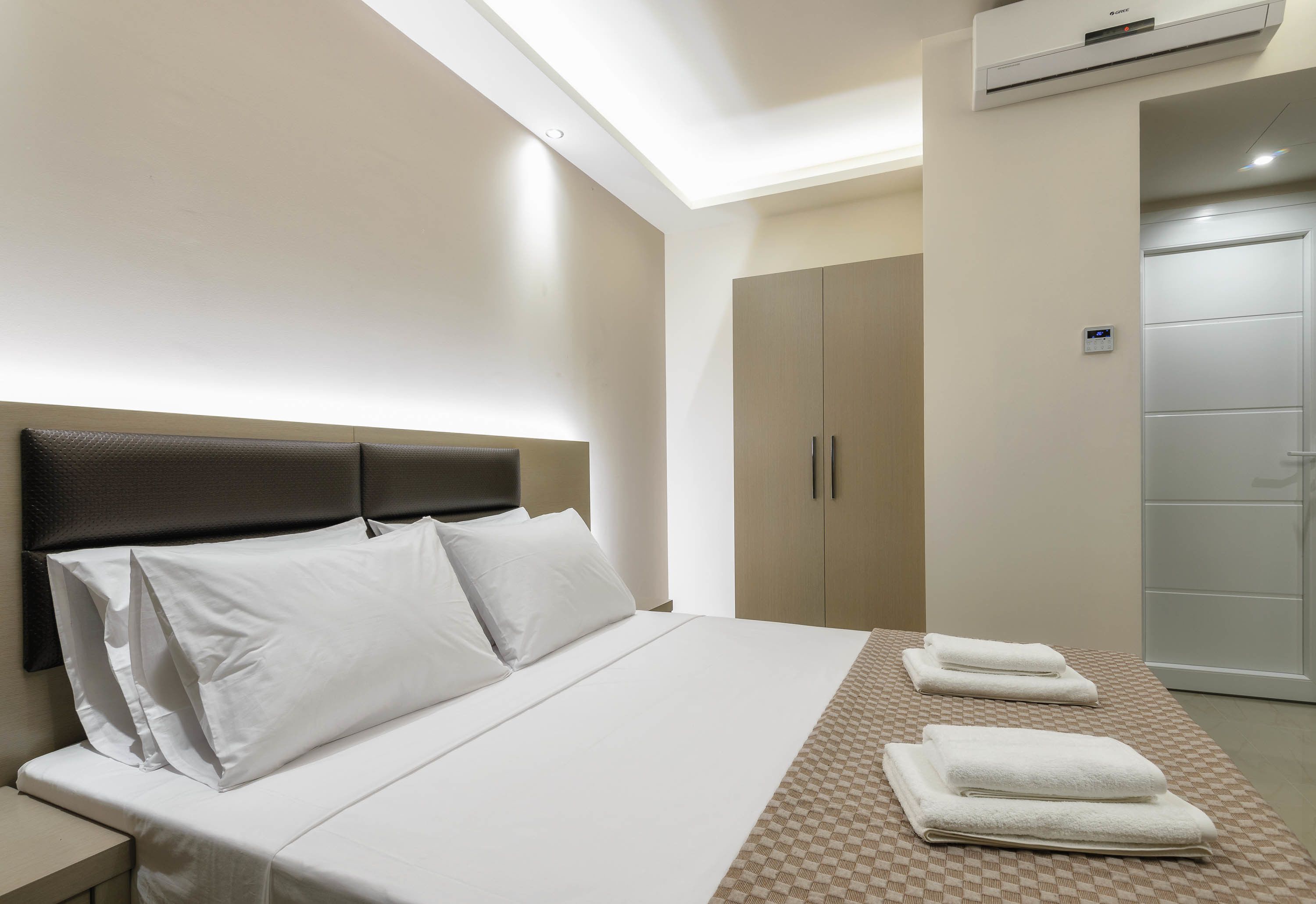 A hotel room with a bed, a nightstand, an air conditioning unit, a wardrobe, and a door.