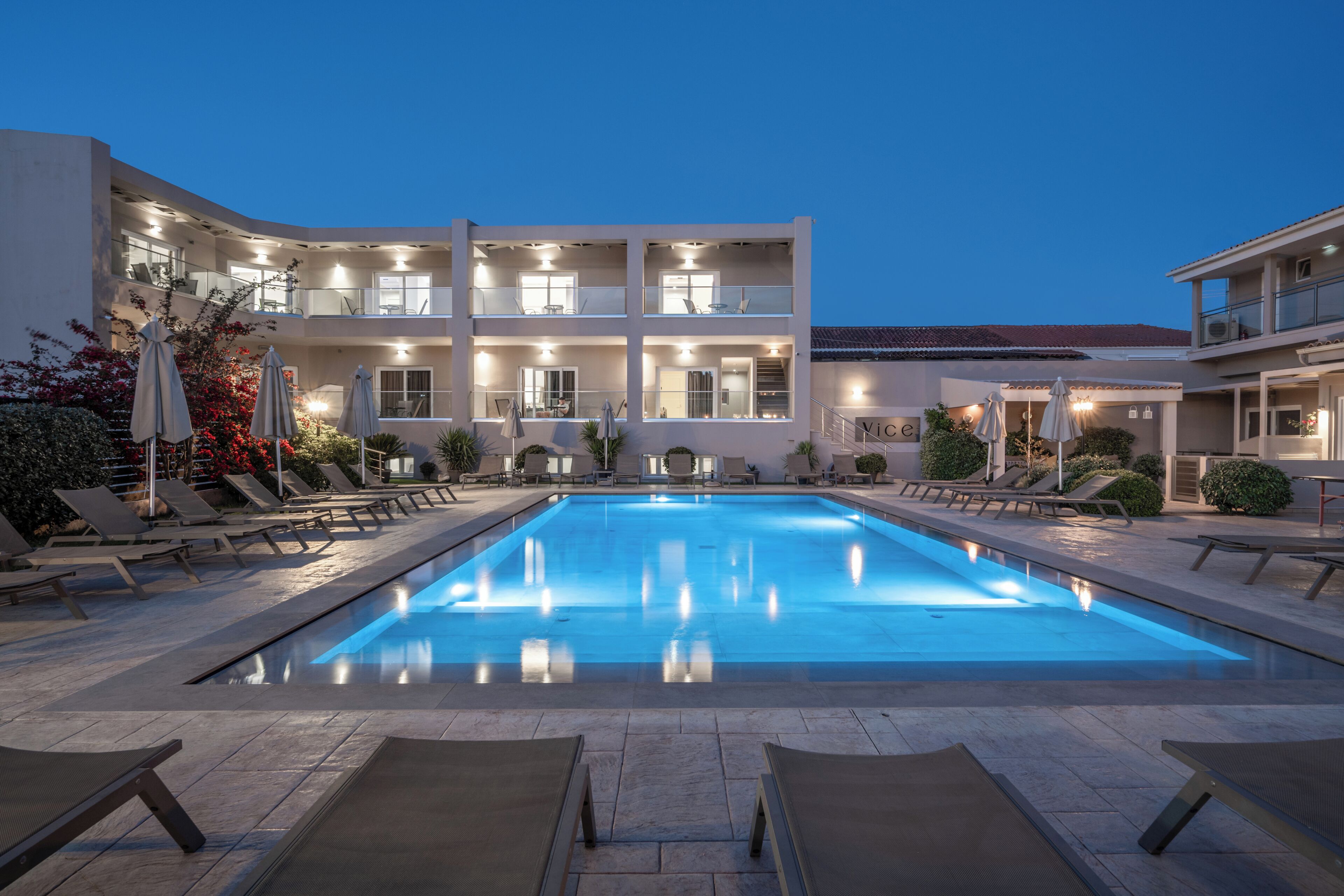 Una piscina con tumbonas, un edificio con varios balcones y un cielo despejado.
