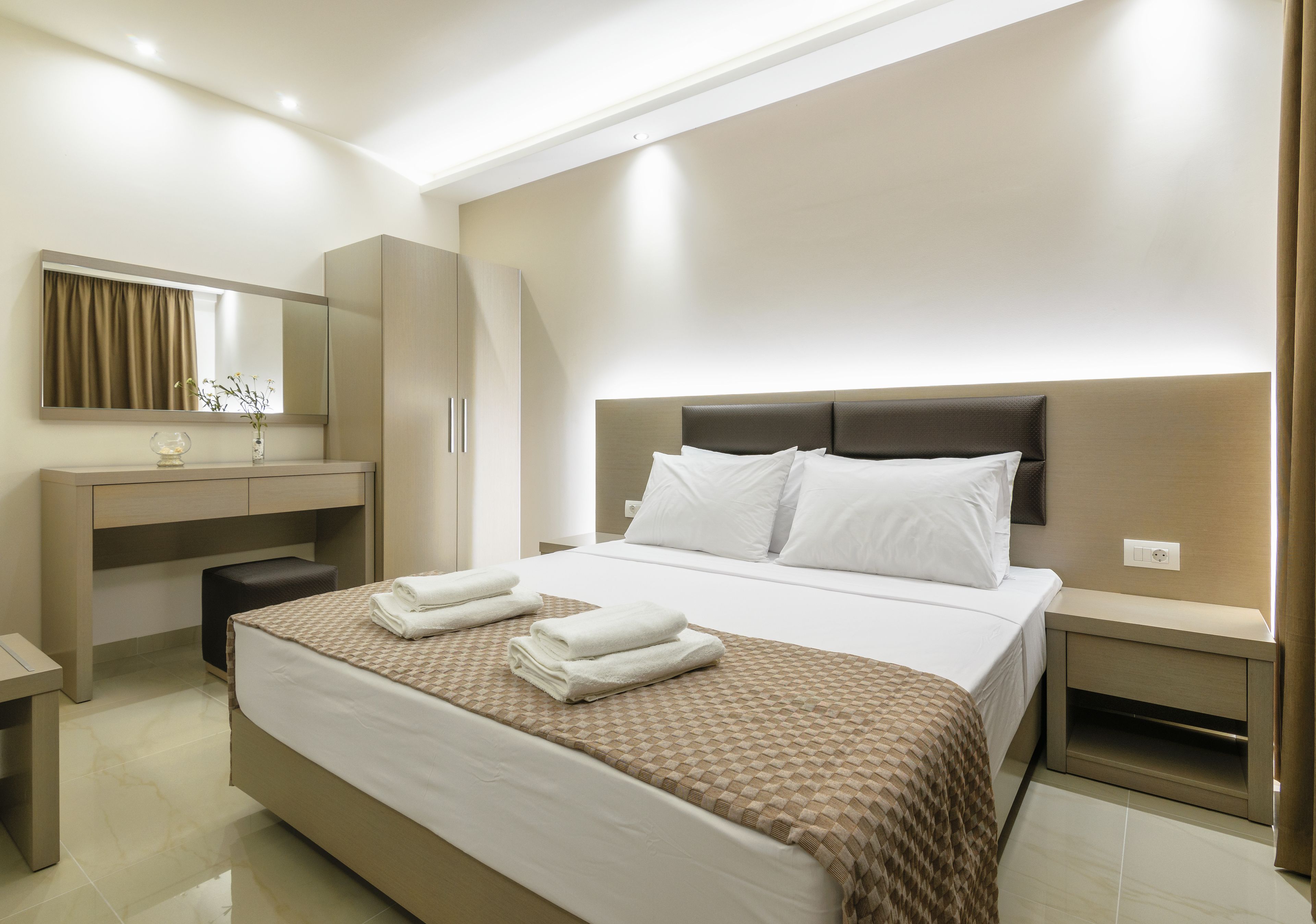 A hotel room with a large bed, a bedside table, a desk with a chair, a mirror, and a wardrobe.