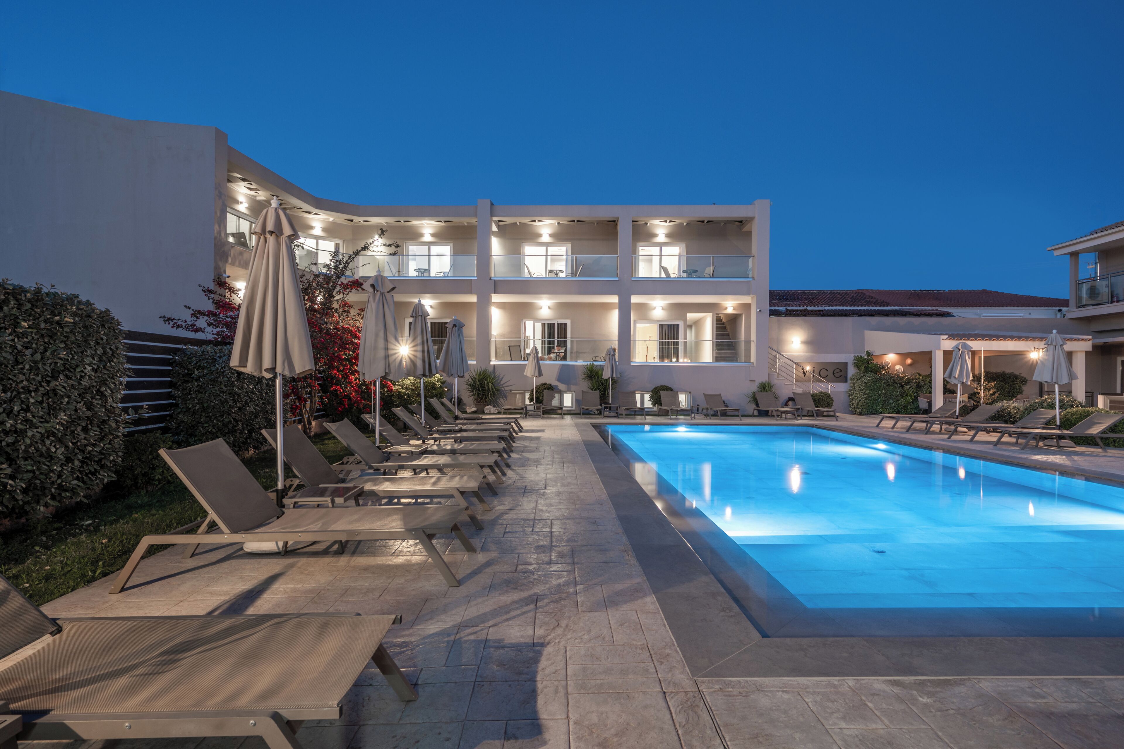 A swimming pool with lounge chairs and umbrellas, a multi-story building with balconies, and a clear evening sky.