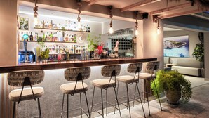 Una zona de bar con mostrador de madera, taburetes y una variedad de botellas y vasos.