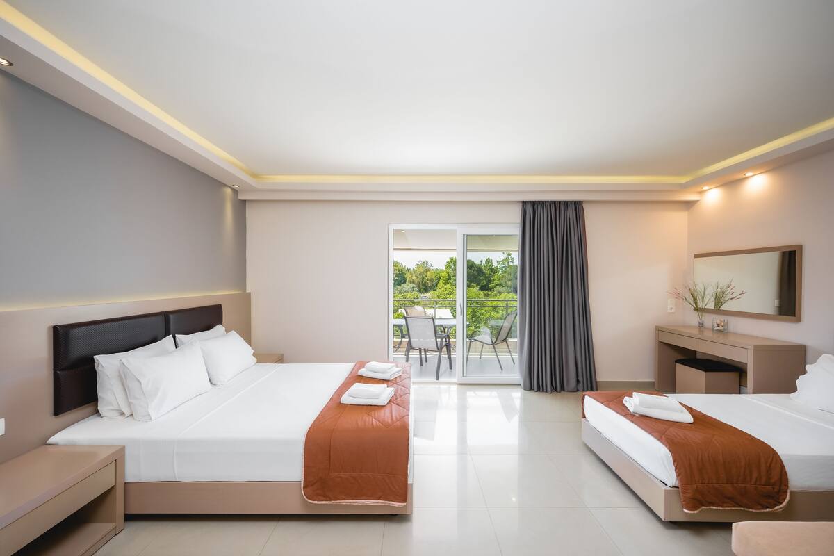 Habitación de hotel con dos camas, un escritorio, una silla y un balcón con vistas a zonas verdes.