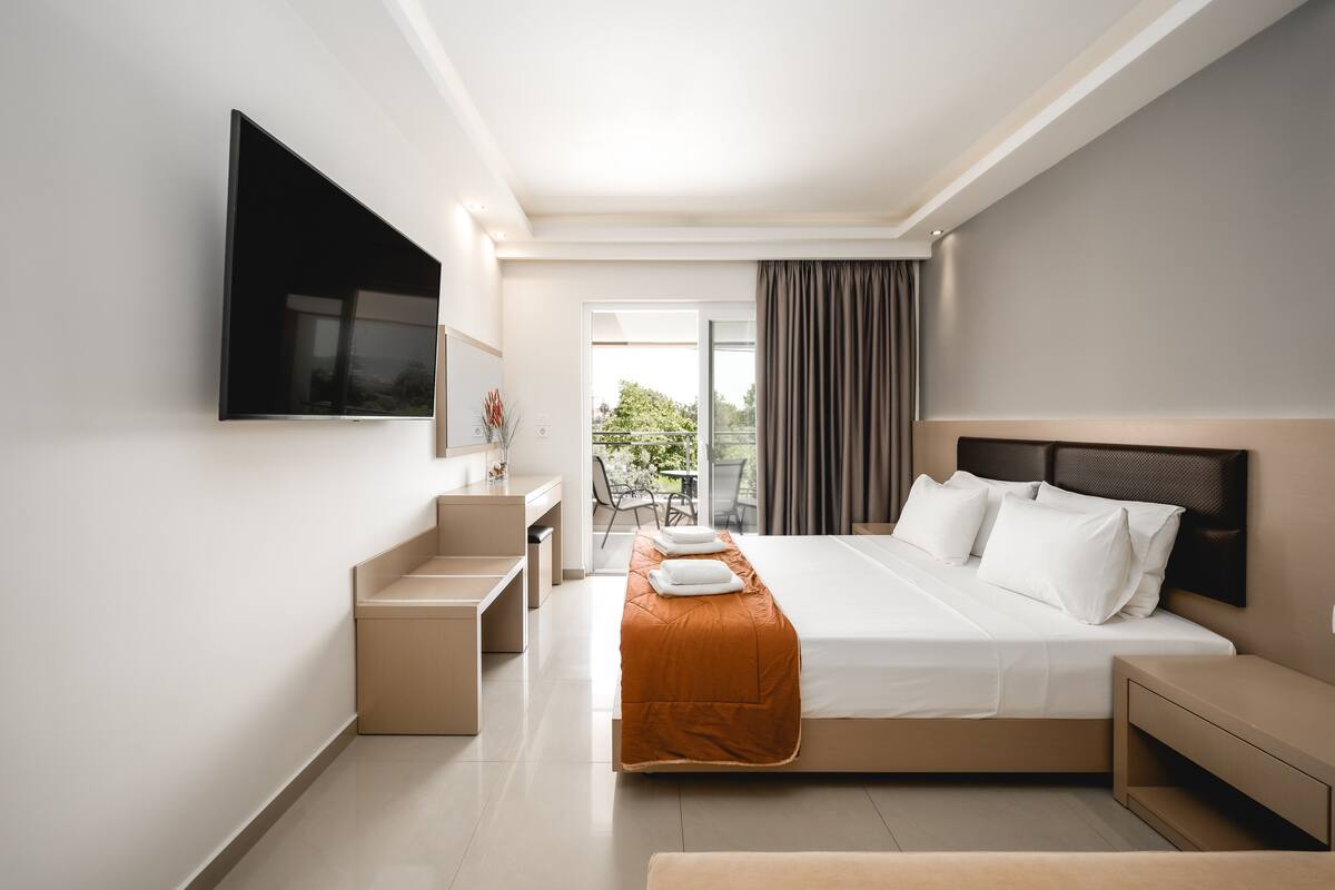 Habitación de hotel moderna con una cama grande, un televisor de pantalla plana montado en la pared, un escritorio pequeño y un balcón con vistas a zonas verdes.