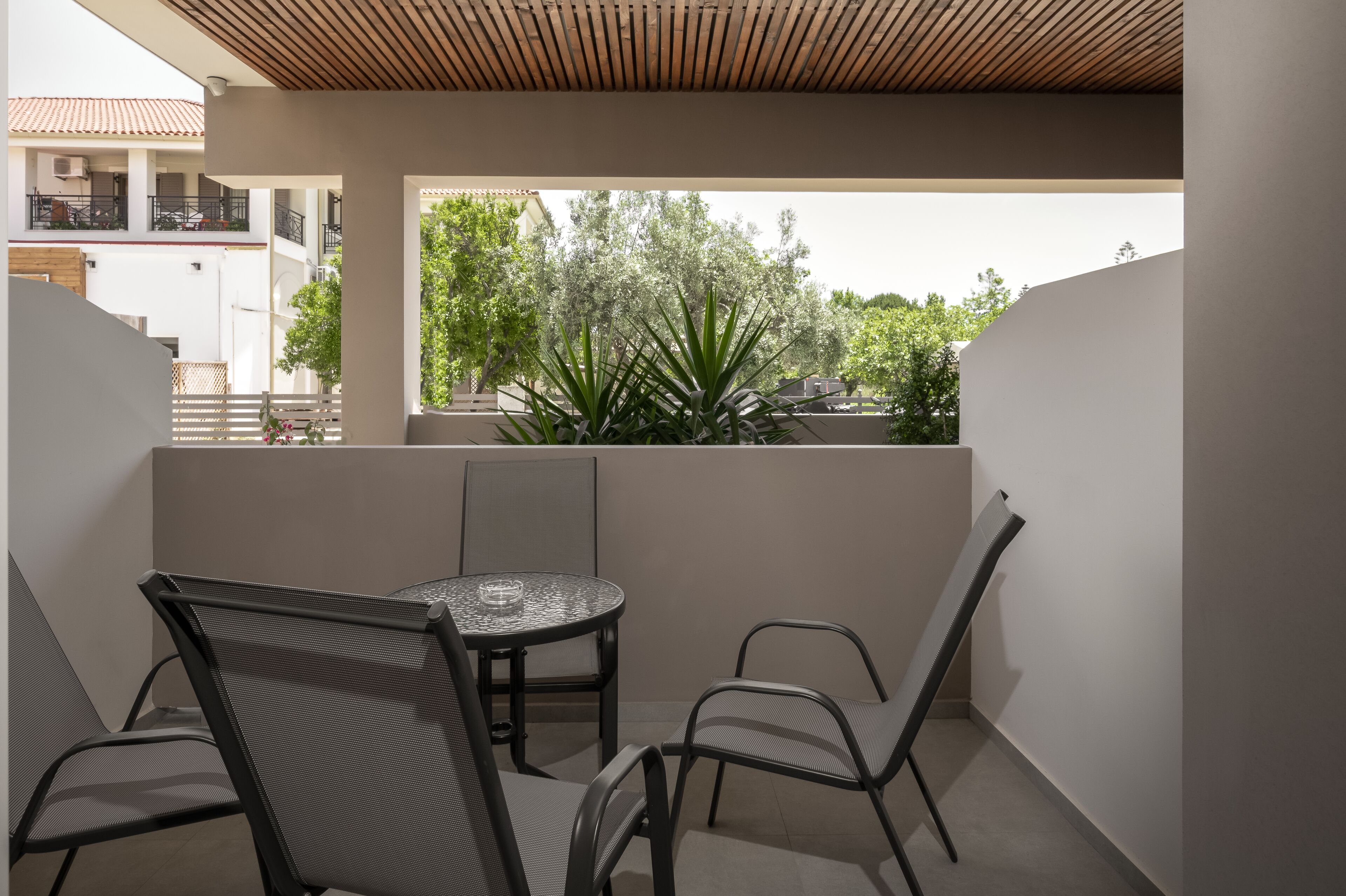 Un balcón con una mesa redonda y cuatro sillas, con vistas a un jardín con plantas altas y un edificio al fondo.