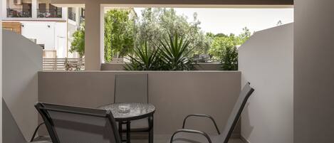 Un balcón con una mesa redonda y cuatro sillas, con vistas a un jardín con plantas altas y un edificio al fondo.