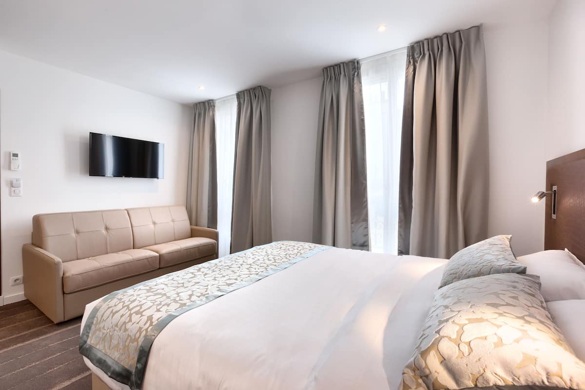 A hotel room with a large bed, a sofa, a flat-screen TV mounted on the wall, and a window with curtains.
