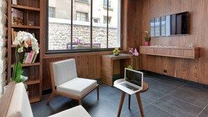 Een moderne kamer met een houten boekenkast, een witte stoel, een bureau met een laptop, en een groot raam met uitzicht op een stenen gebouw.