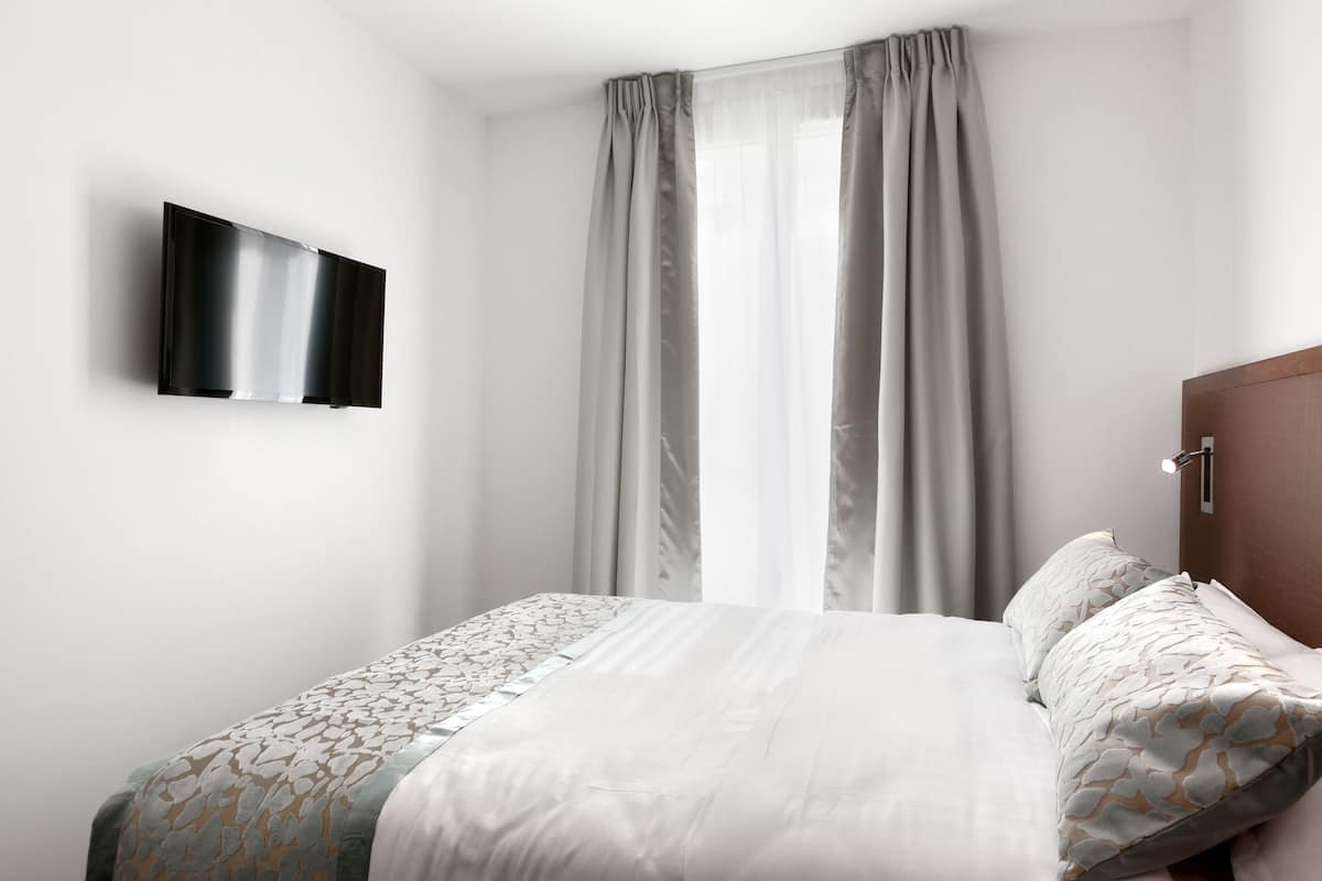 A hotel room with a bed, a flat-screen TV mounted on the wall, and a window with curtains.