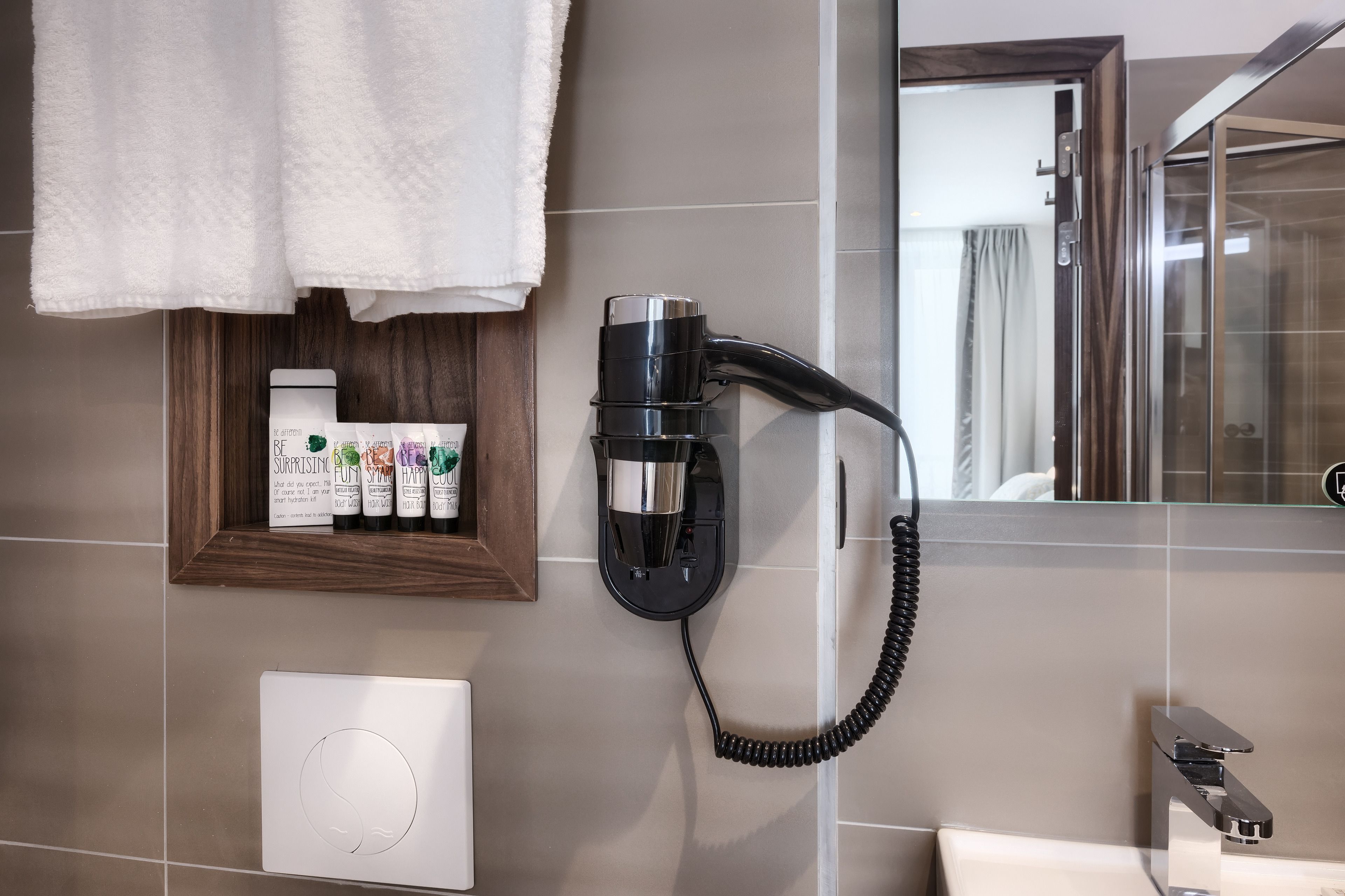 A bathroom with a hairdryer mounted on the wall, a framed shelf with toiletries, and a mirror reflecting a shower area.